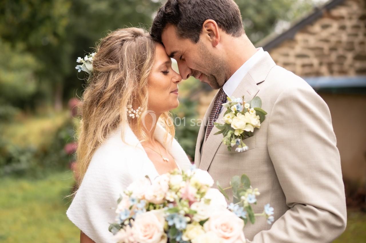 Un couple en tenue de mariage se tient face à face dans un jardin, tenant un bouquet de fleurs pastel.