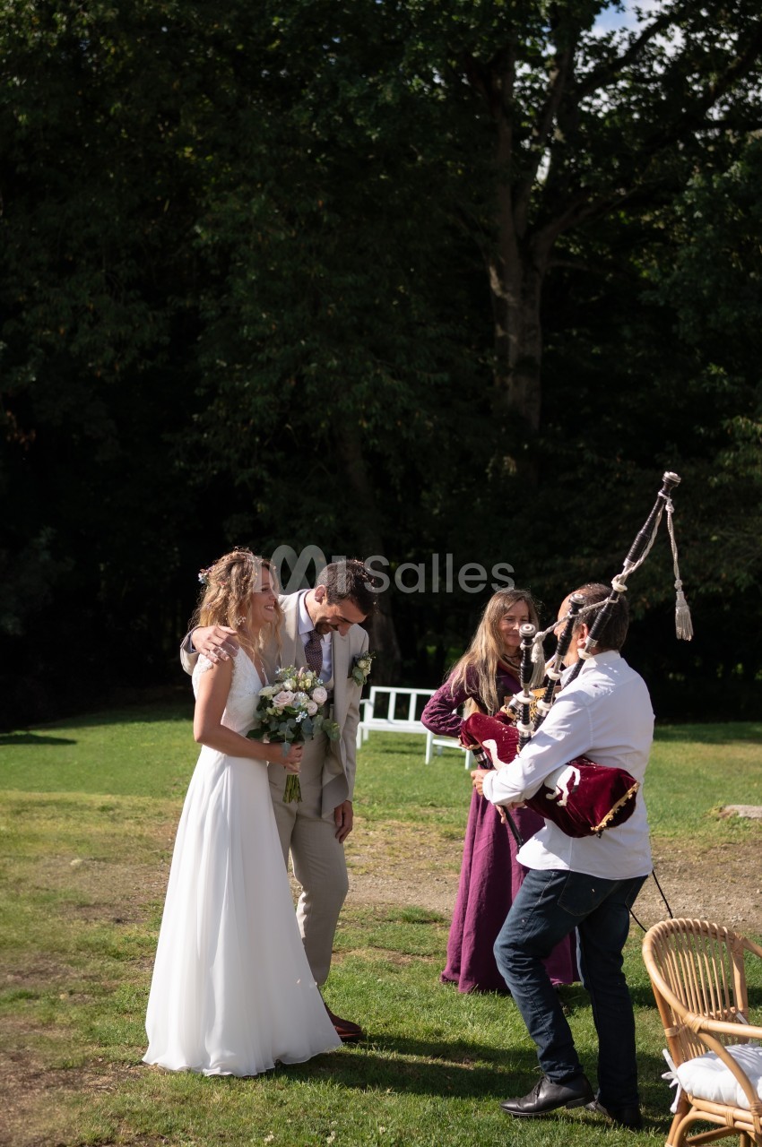 Un couple de mariés sourit en extérieur, accompagné de musiciens jouant de la cornemuse dans un cadre verdoyant.