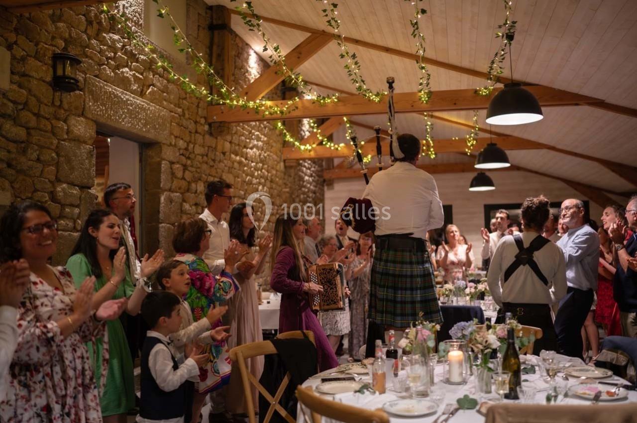 Un joueur de cornemuse en kilt joue devant des invités debout dans une salle de réception décorée de guirlandes lumineuses.