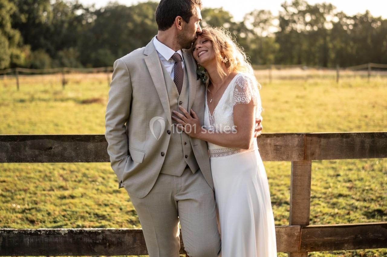 Un couple en tenue de mariage s'embrasse et sourit devant une clôture en bois dans un champ ensoleillé.