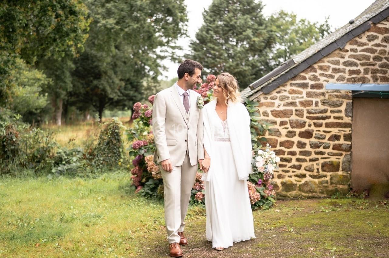 Un couple en tenue de mariage marche côte à côte dans un jardin près d'un bâtiment en pierre.