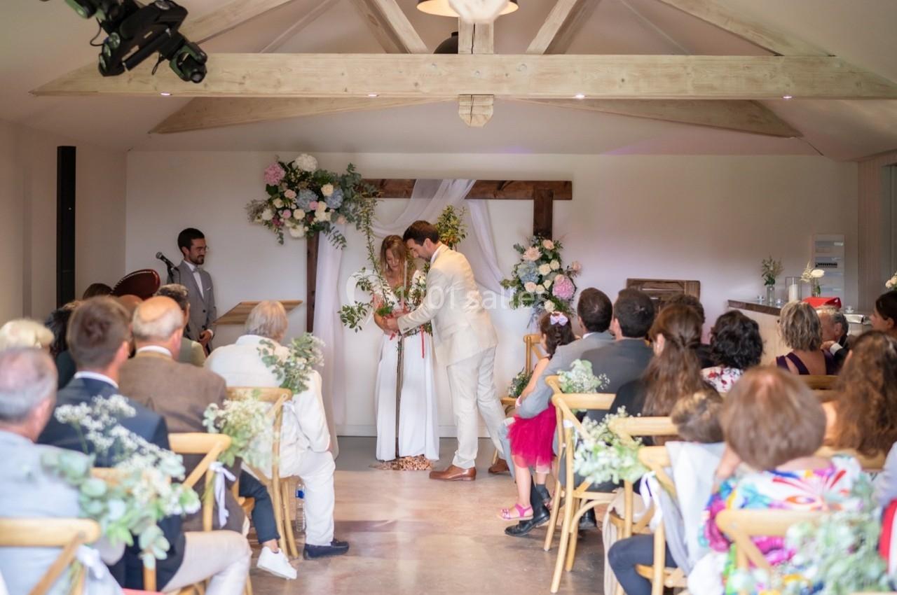 Cérémonie de mariage dans une salle lumineuse avec des invités assis et un couple échangeant leurs vœux devant une arche…