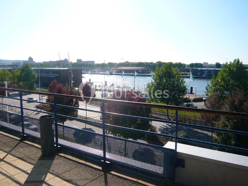 Vue sur un fleuve bordé d'arbres, avec des bateaux amarrés et des bâtiments en arrière-plan, depuis une terrasse.