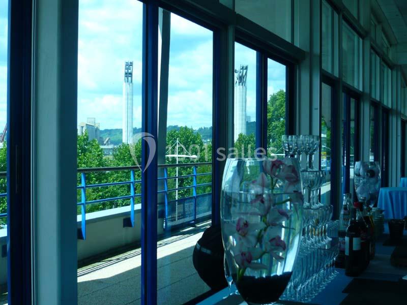 Vue d'une salle vitrée donnant sur une terrasse avec des arbres, des verres alignés et un vase contenant des fleurs.
