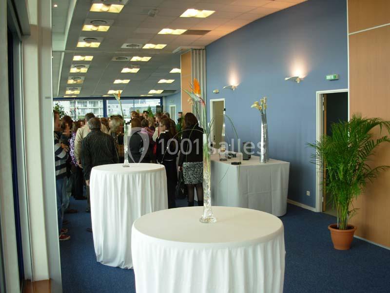 Salle de réception lumineuse avec tables hautes décorées de fleurs et un groupe de personnes en arrière-plan.