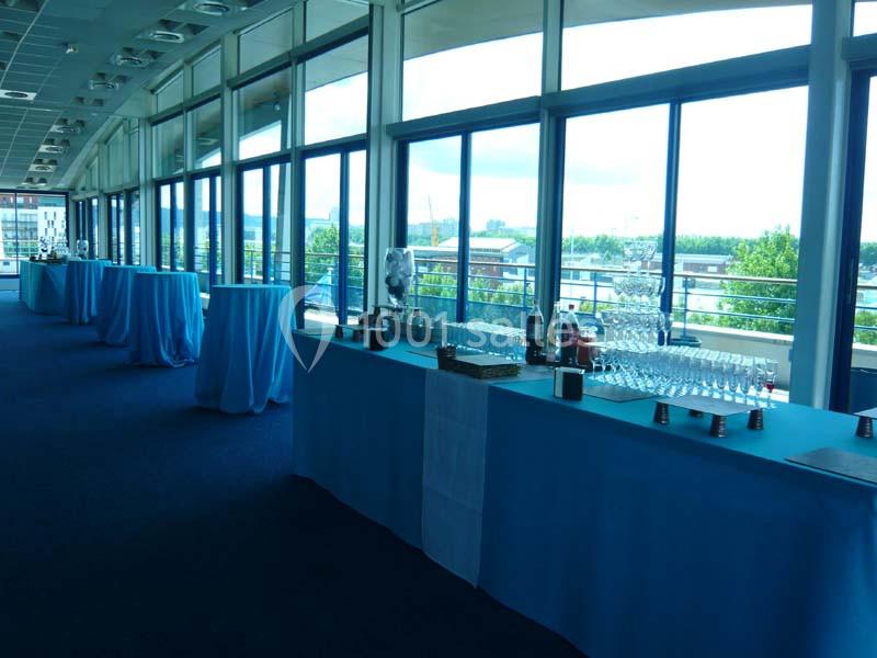 Tables dressées avec nappes bleues, verres et accessoires, alignées dans une salle lumineuse avec de grandes baies vitrées.
