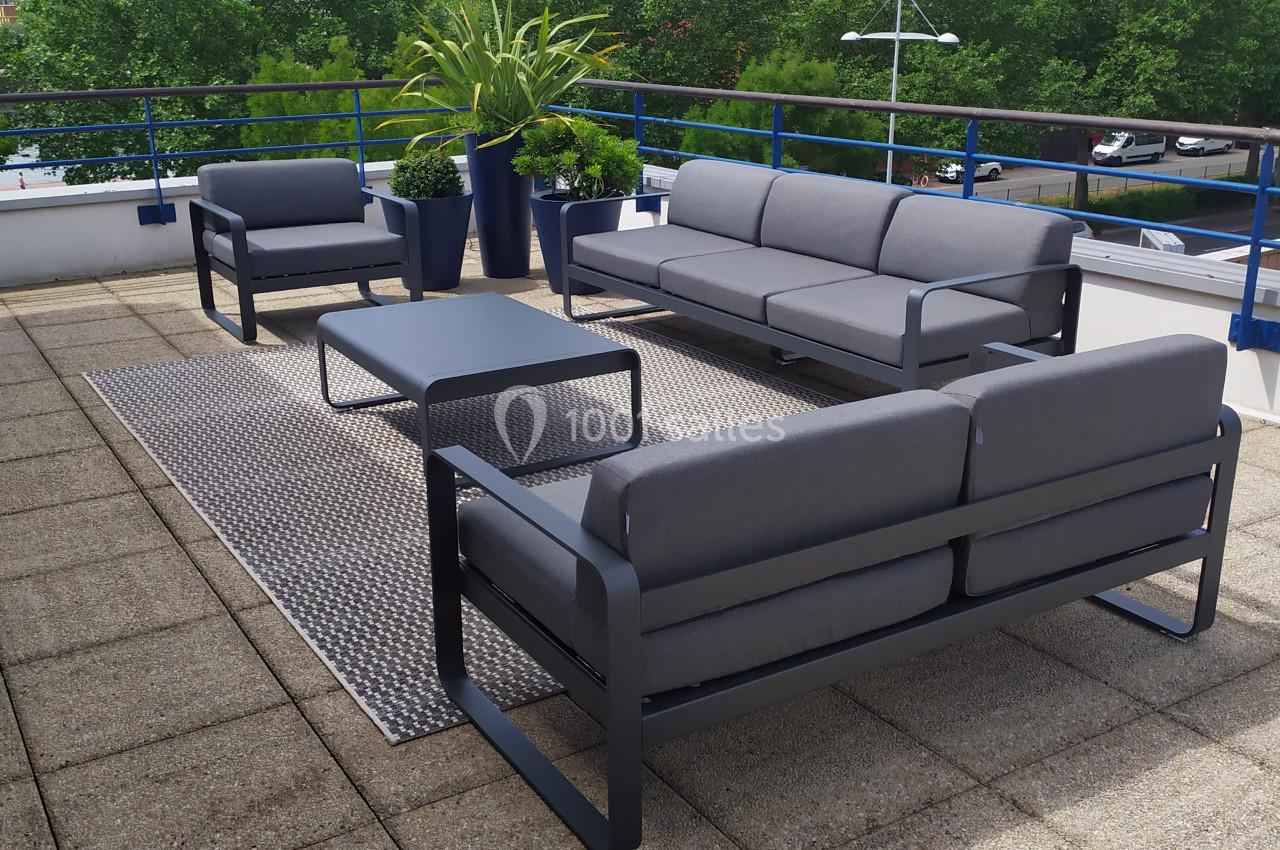 Salon de jardin moderne avec canapés gris, table basse et plantes, installé sur une terrasse ensoleillée avec vue arborée.