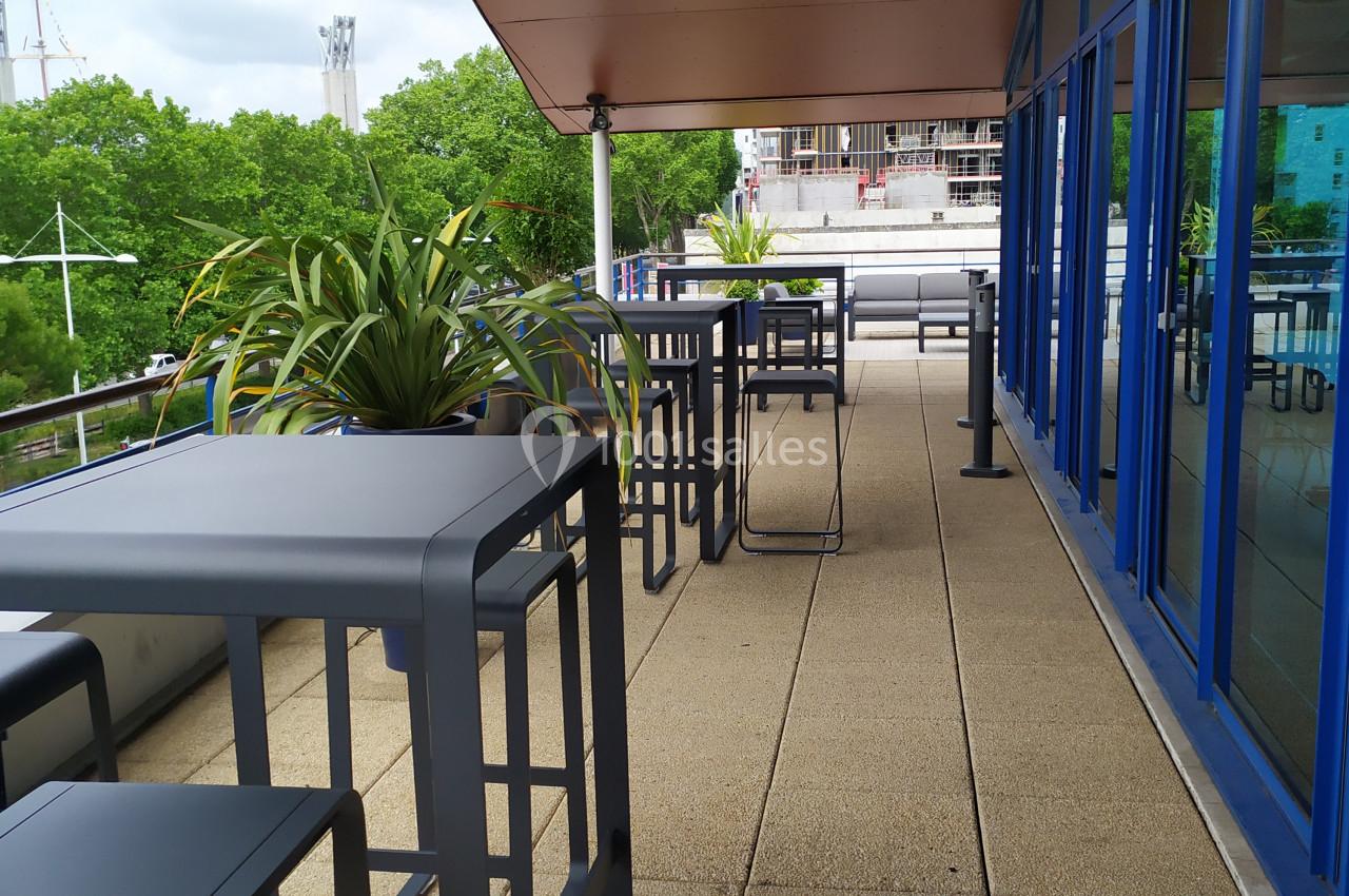 Terrasse avec tables et tabourets hauts en métal, entourée de plantes, donnant sur un paysage urbain et arboré.