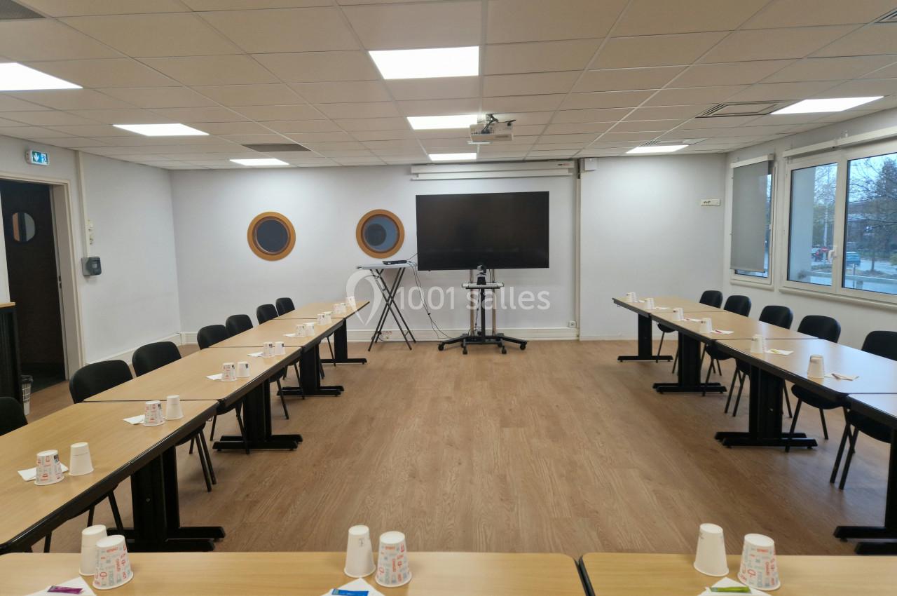 Salle de réunion équipée de tables disposées en U, avec gobelets et papiers, écran et fenêtres donnant sur l'extérieur.