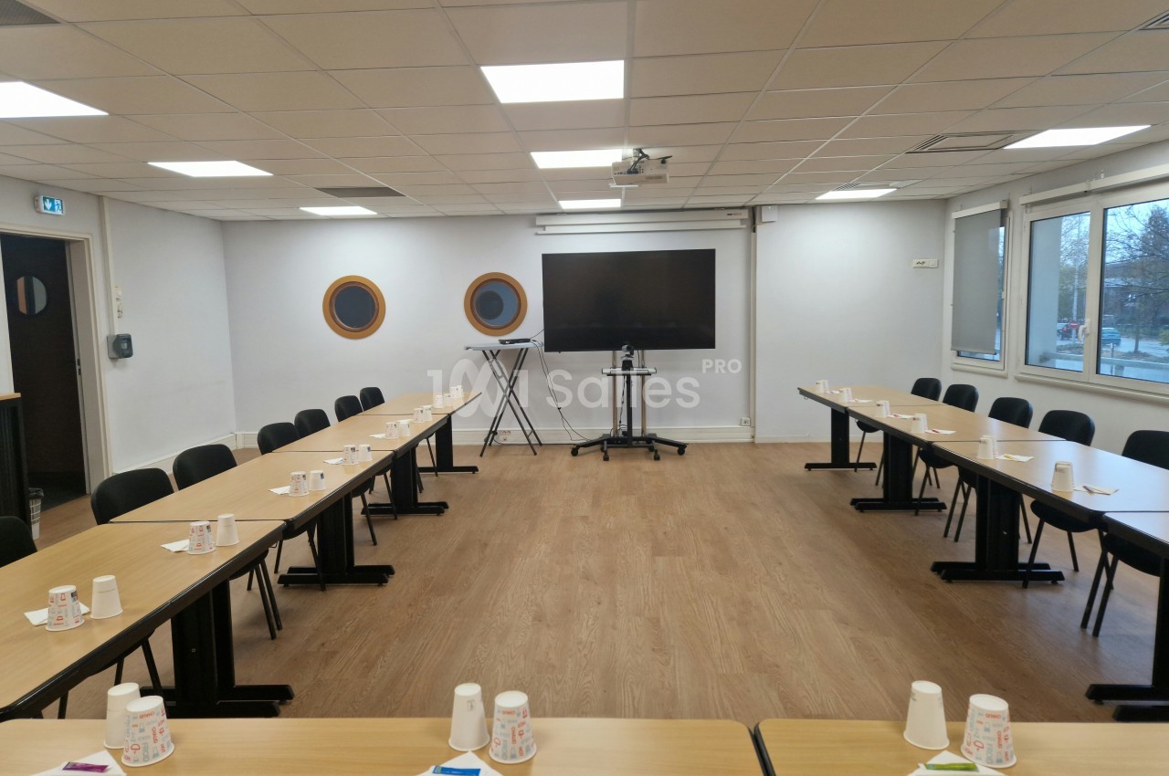 Salle de réunion équipée de tables disposées en U, avec gobelets et papiers, écran et fenêtres donnant sur l'extérieur.