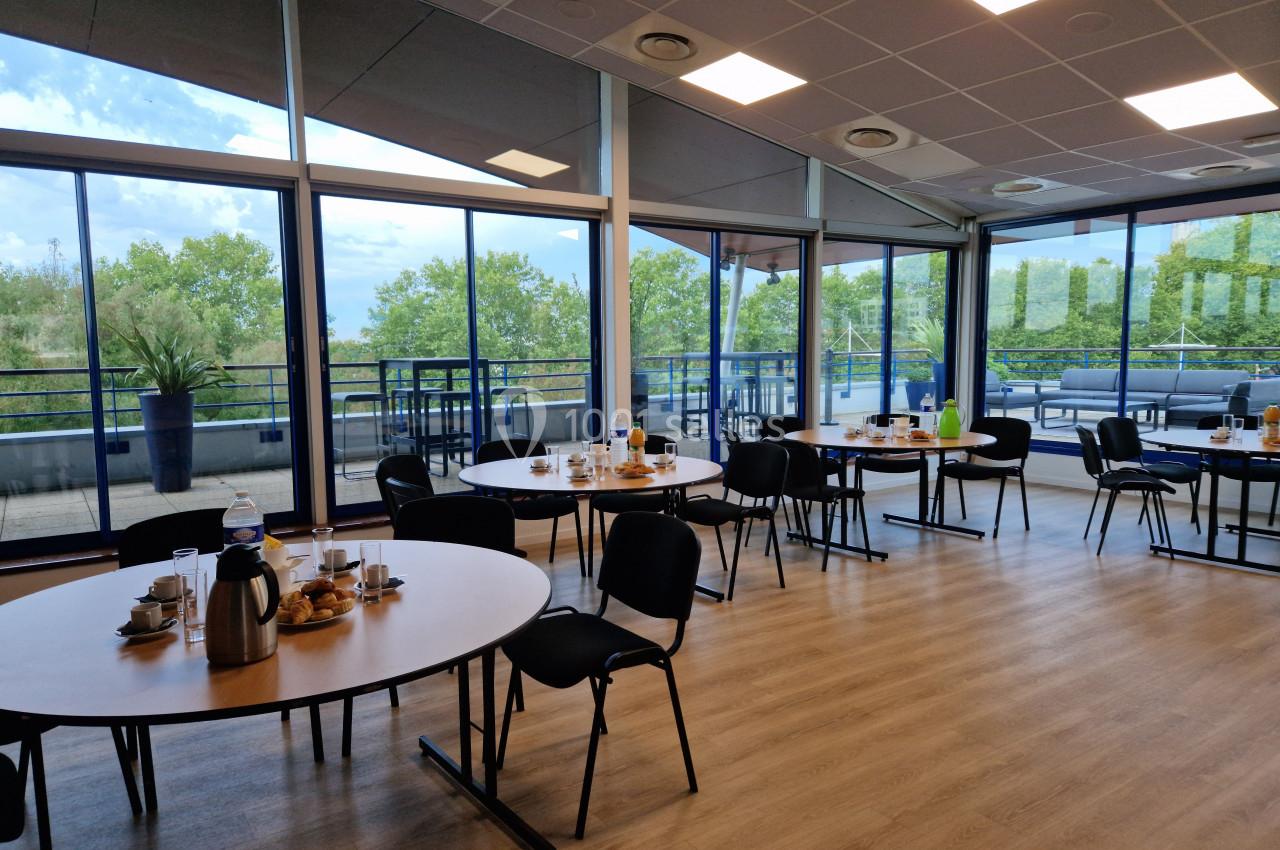 Salle lumineuse avec tables rondes dressées pour un petit-déjeuner, grandes baies vitrées donnant sur une terrasse arborée.