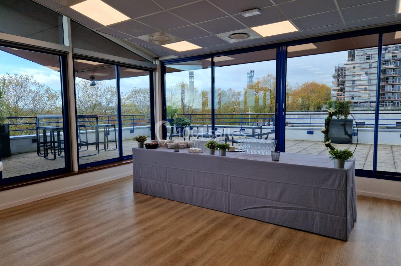Salle lumineuse avec une table recouverte d'une nappe grise, vue sur une terrasse et des arbres à travers de grandes baies…