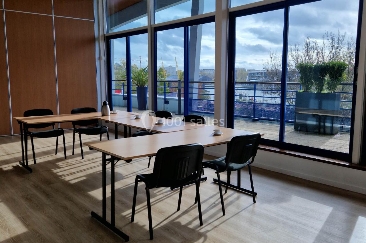 Salle de réunion lumineuse avec grandes baies vitrées, tables en bois, chaises noires et vue sur une terrasse arborée.