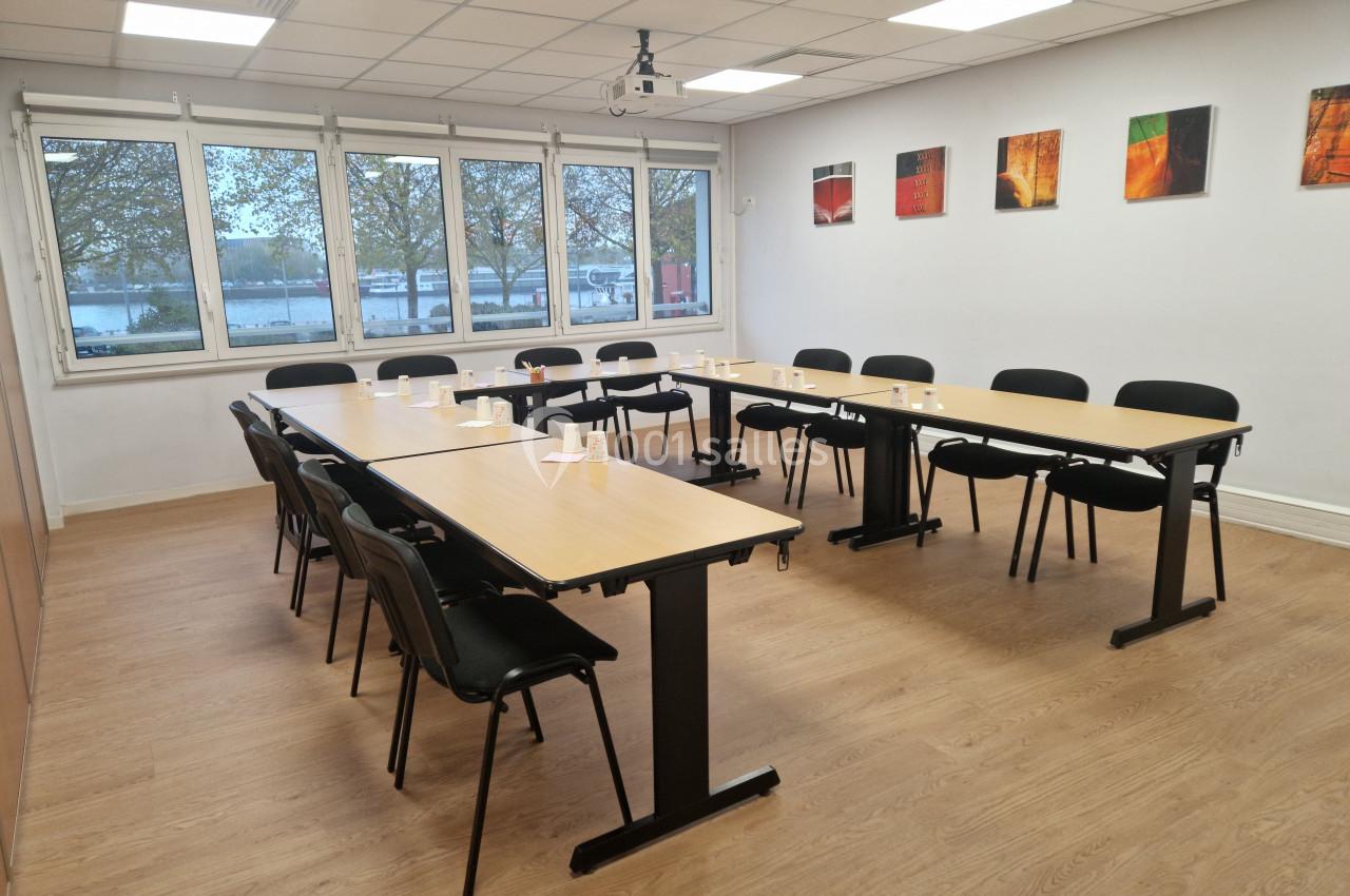 Salle de réunion lumineuse avec tables en U, chaises noires, parquet clair et fenêtres donnant sur un extérieur arboré.