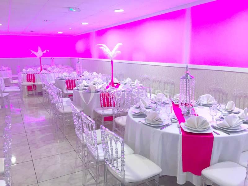 Salle de réception décorée avec des tables rondes, nappes blanches, chemins de table roses et éclairage rose.