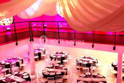 Salle de réception décorée avec des tables rondes, nappes blanches, chaises transparentes et centres de table fleuris.