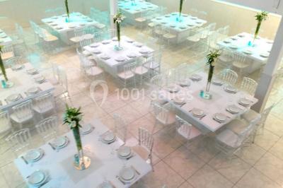 Salle de réception décorée avec des tables rondes, nappes blanches, chaises transparentes et centres de table fleuris.