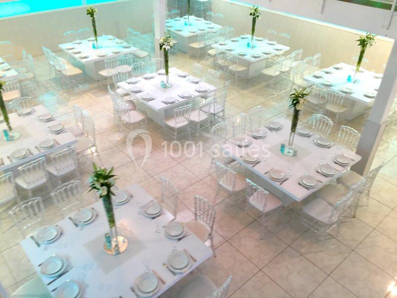 Salle de réception lumineuse avec des tables carrées dressées, décorées de vases hauts contenant des fleurs blanches.