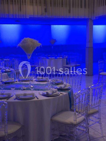 Salle de réception avec tables rondes dressées, nappes blanches, chaises transparentes et éclairage bleu tamisé.