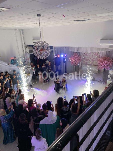 Des invités entourent un couple assis au centre d'une salle décorée, avec des étincelles et de la fumée au sol.