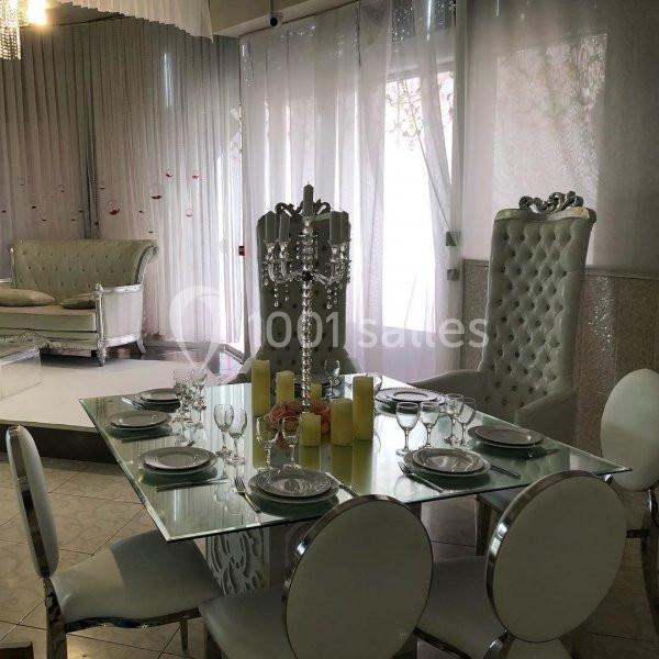 Table en verre dressée avec vaisselle et bougies jaunes, entourée de chaises blanches, dans un salon élégant avec rideaux.