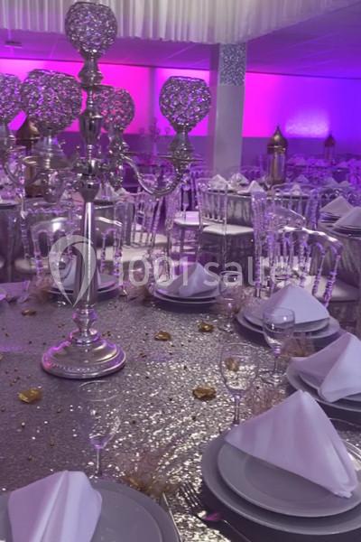 Salle de réception décorée avec des tables dressées, nappes scintillantes, chandeliers argentés et éclairage rose.