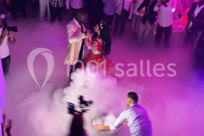 Salle de réception décorée avec des tables rondes, nappes blanches, chaises transparentes et centres de table fleuris.