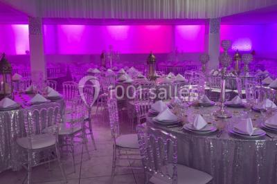 Salle de réception décorée avec des tables rondes, nappes blanches, chaises transparentes et centres de table fleuris.