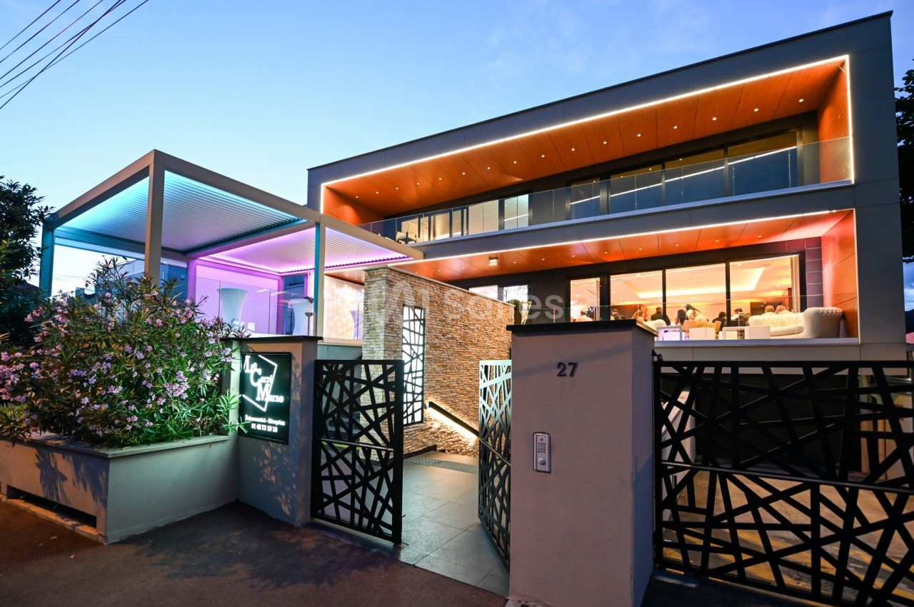 Façade moderne d'un bâtiment éclairé par des lumières colorées au crépuscule, avec une entrée design et végétation.