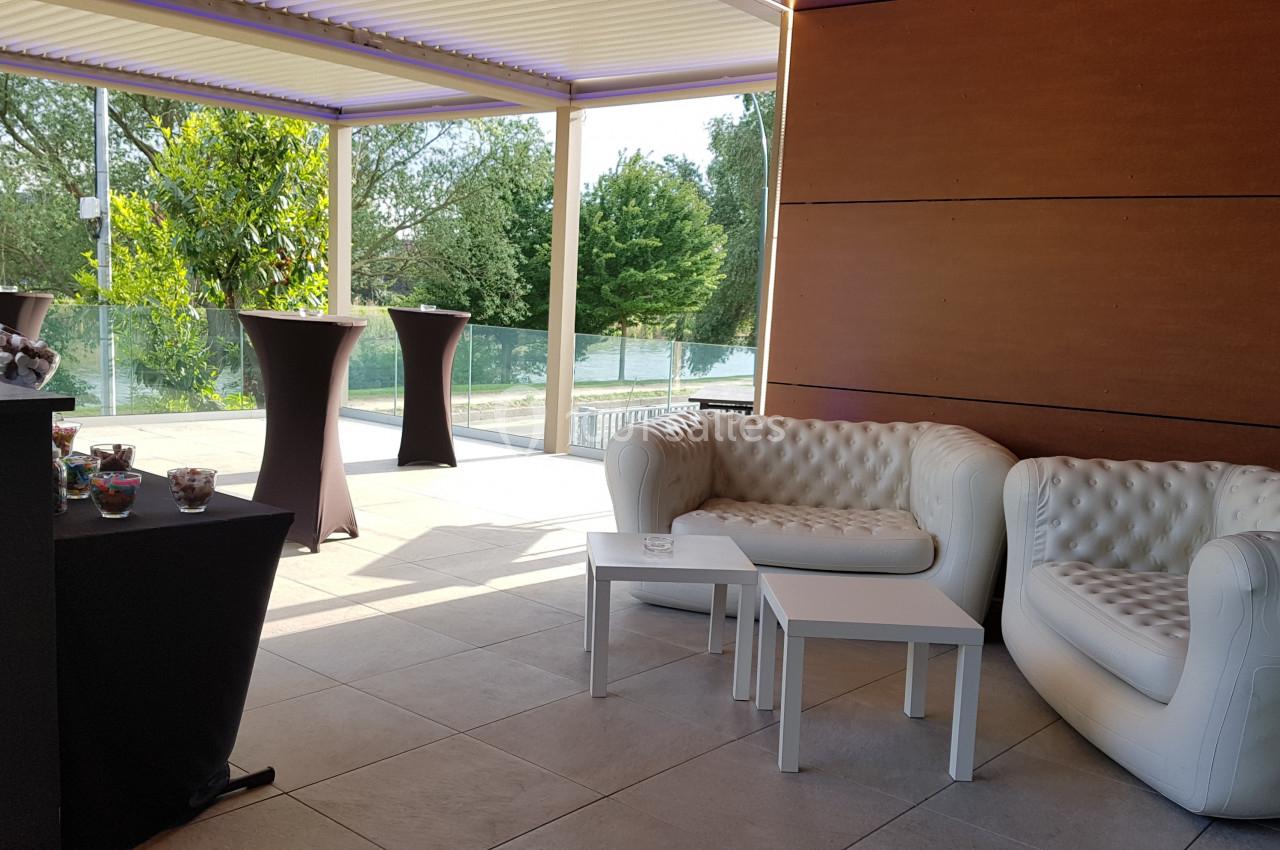 Salon extérieur avec deux canapés blancs, tables basses et vue sur une terrasse arborée avec des tables hautes.