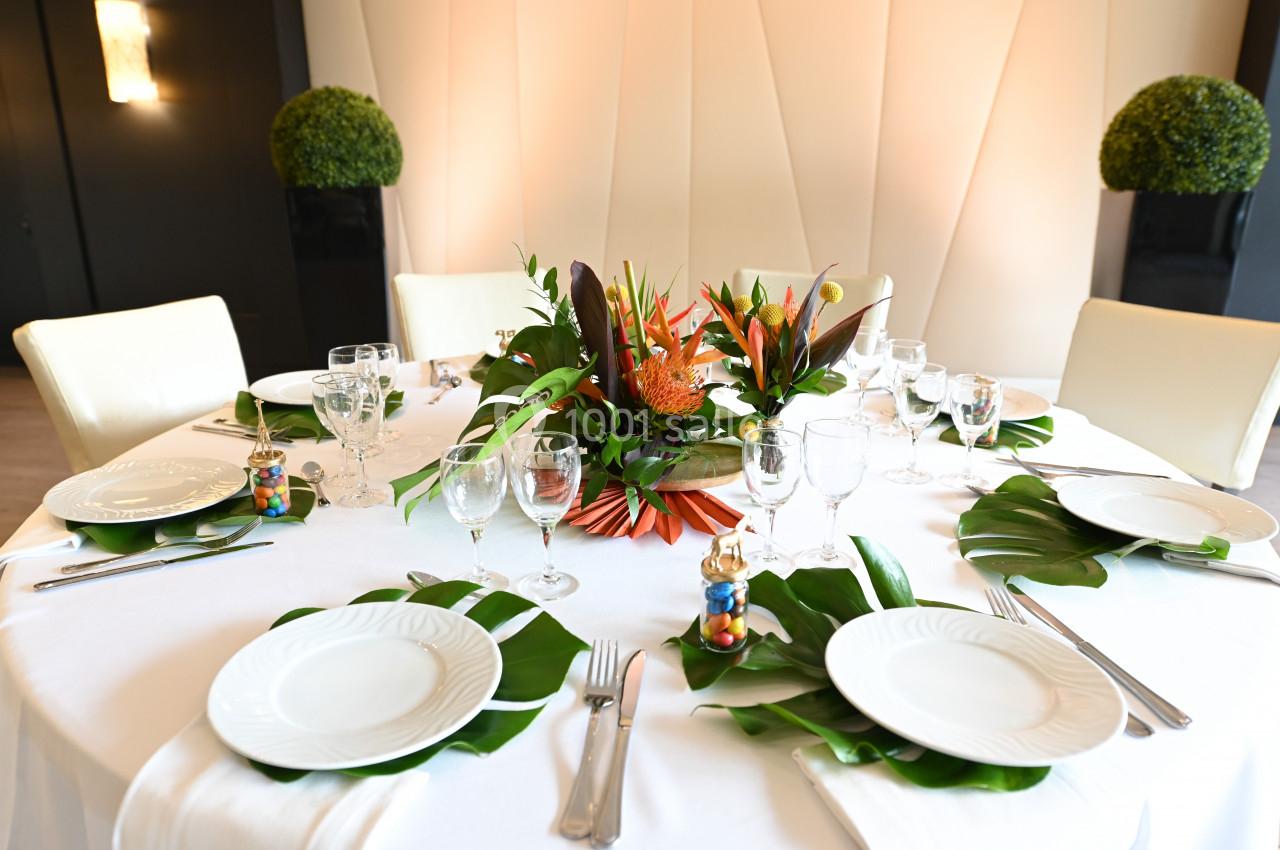 Table dressée avec une nappe blanche, vaisselle élégante, décoration florale exotique et feuilles vertes en centre de table.