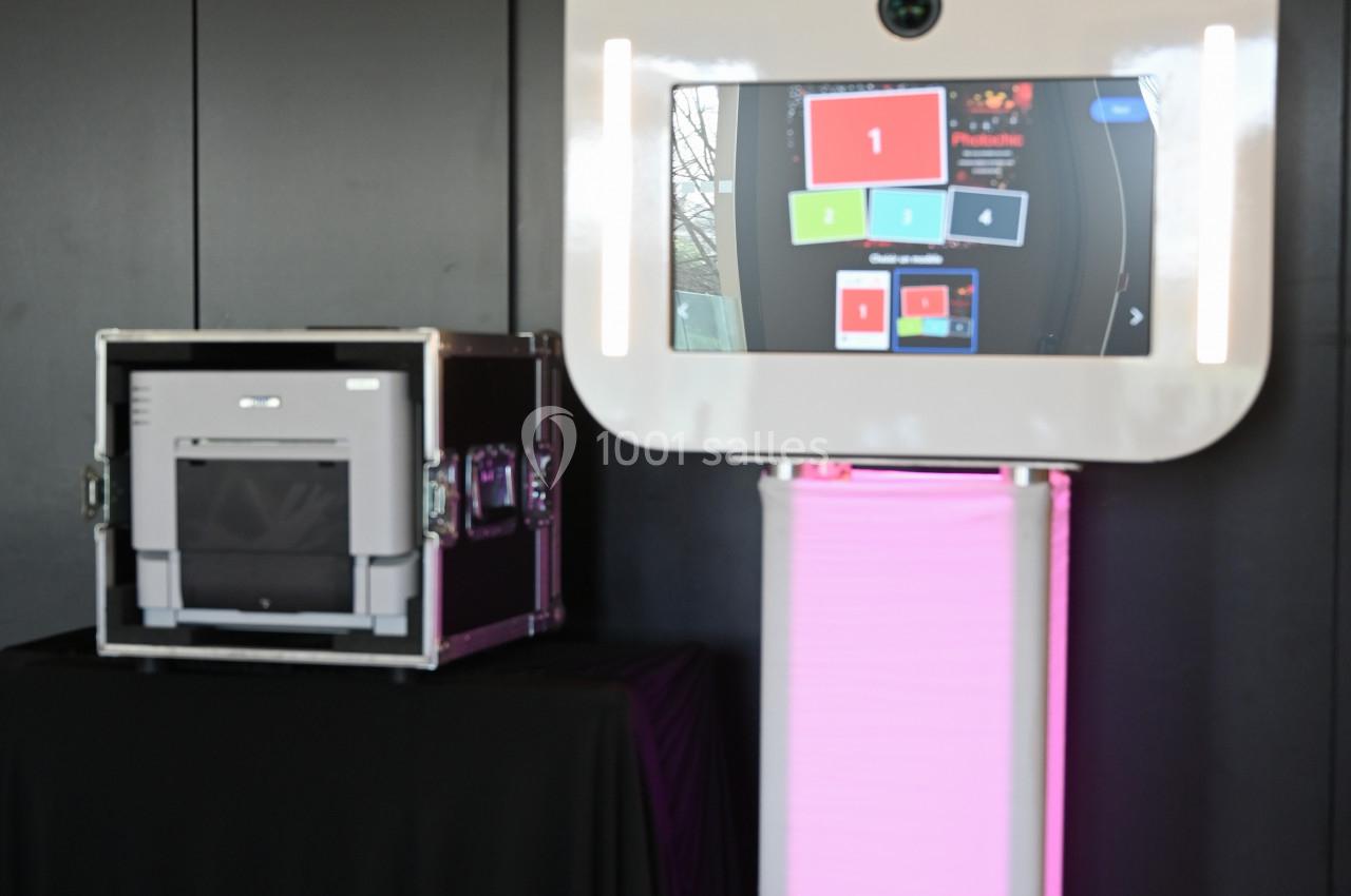 Borne photo interactive sur pied avec écran lumineux, placée à côté d'une mallette noire dans un environnement intérieur.