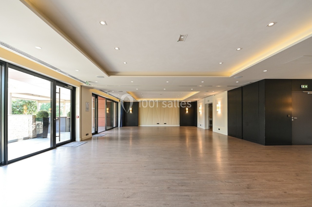 Salle vide avec parquet clair, murs neutres, éclairage encastré et grandes baies vitrées donnant sur l'extérieur.