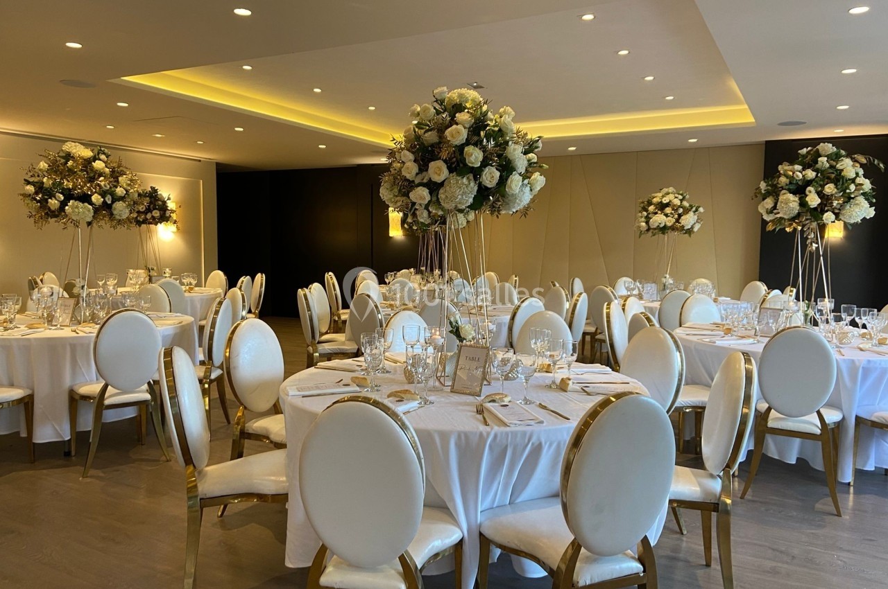 Salle de réception élégante avec tables rondes dressées, décorations florales blanches et chaises aux dossiers arrondis.