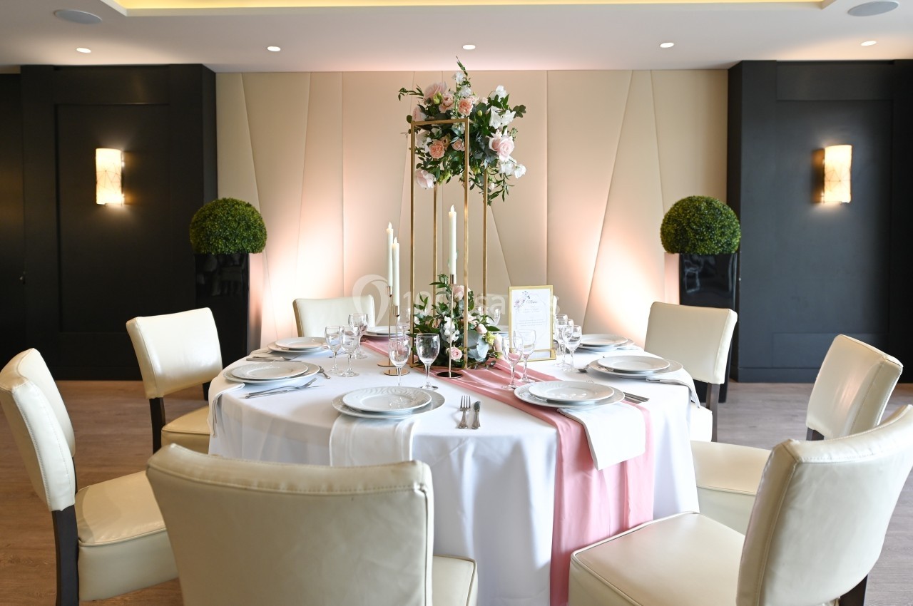 Table ronde élégamment dressée avec nappes blanches, décoration florale et bougies, entourée de chaises rembourrées.