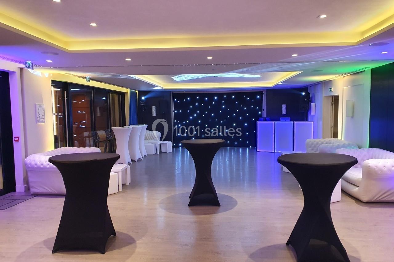 Salle de réception moderne avec tables hautes noires, canapés blancs et éclairage LED coloré.