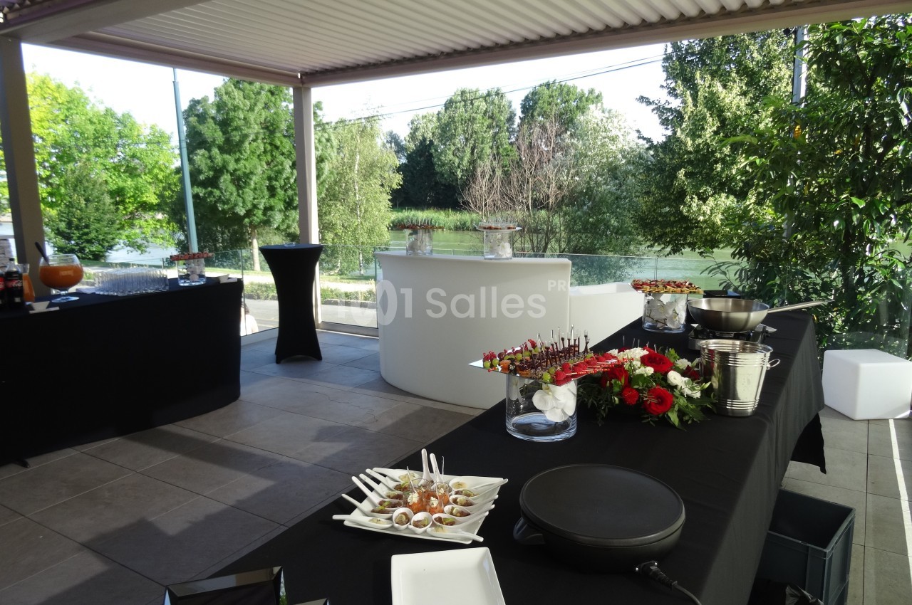 Buffet extérieur avec tables noires garnies de fleurs, amuse-bouches et boissons, sous une pergola près d'un jardin.
