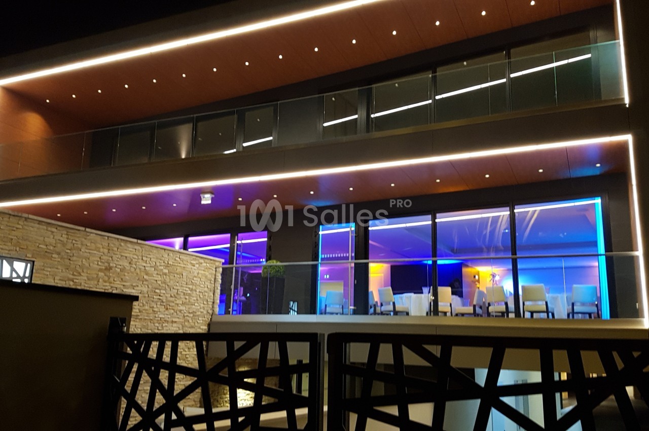 Façade moderne d'un bâtiment éclairé de nuit avec des lumières colorées et une terrasse vitrée.