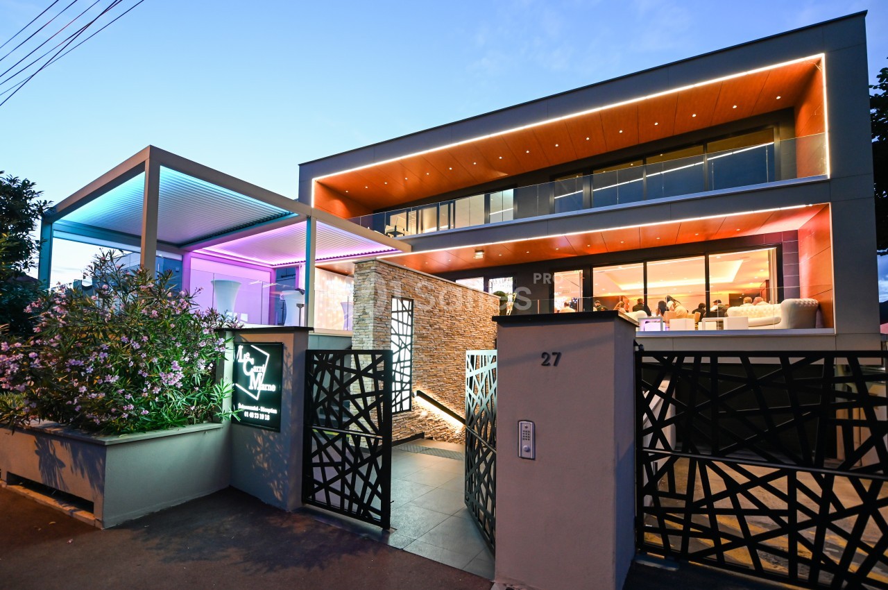 Façade moderne d'un bâtiment éclairé par des lumières colorées au crépuscule, avec une entrée design et végétation.