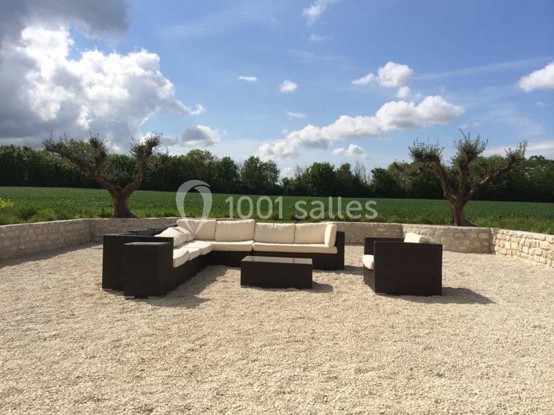 Salon de jardin en résine tressée avec coussins beiges, installé sur une terrasse en gravier, entourée de verdure.