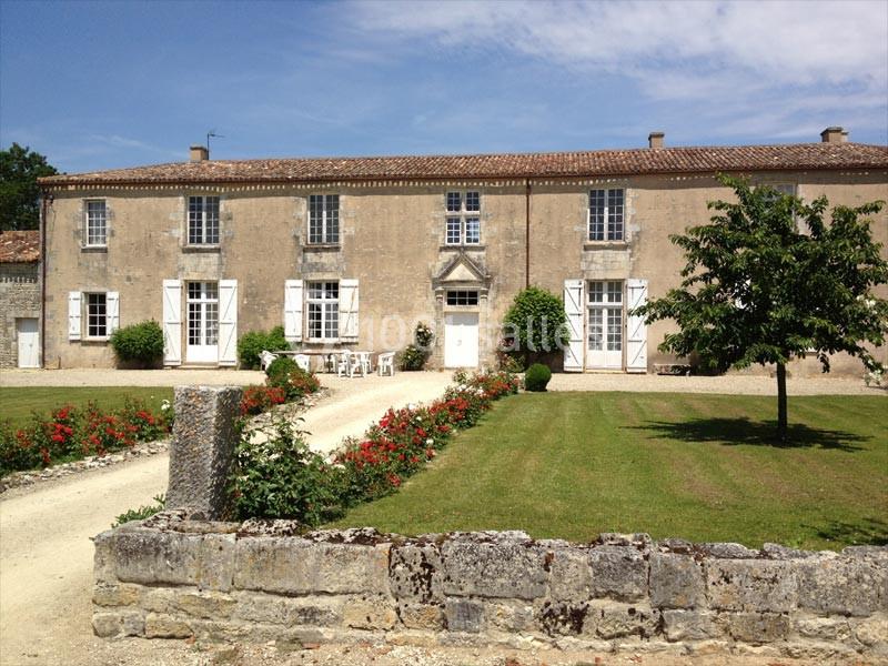 Façade d'une grande maison en pierre avec volets blancs, jardin fleuri et allée gravillonnée sous un ciel dégagé.