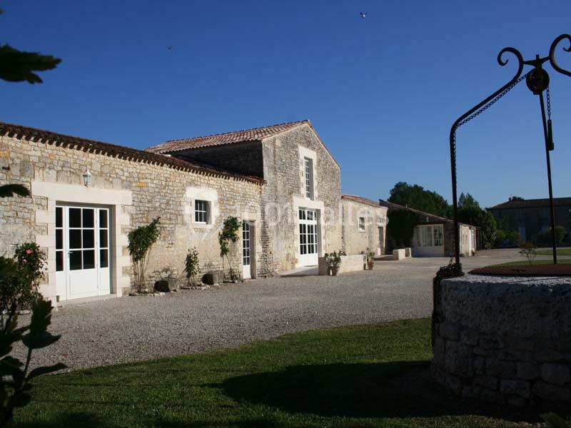 Bâtiment en pierre avec des portes vitrées, entouré de végétation et d'une cour gravillonnée sous un ciel bleu.