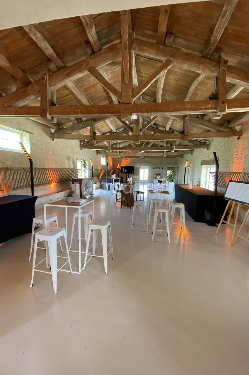Grande salle avec charpente en bois apparente, tables hautes blanches et décoration lumineuse chaleureuse.