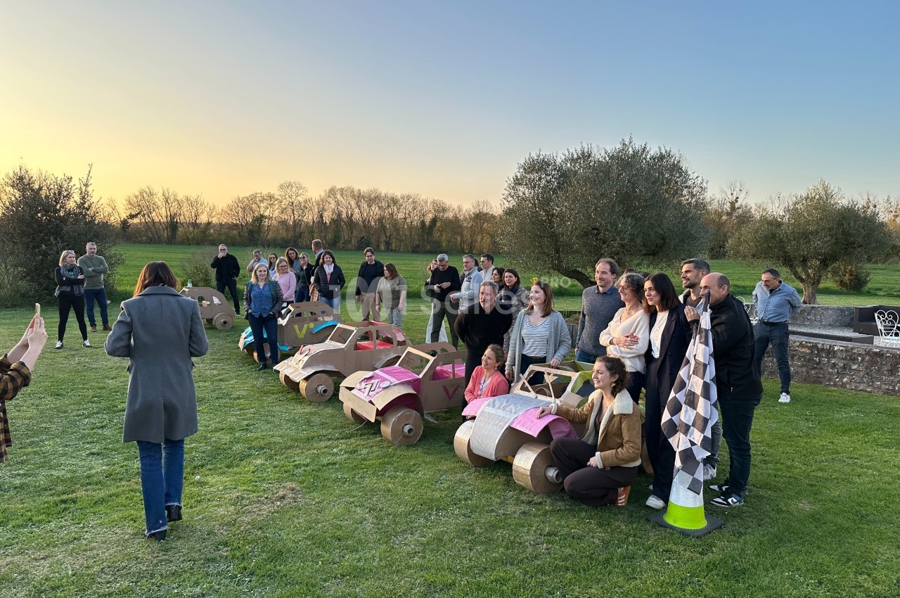 Un groupe de personnes posant avec des voitures en carton sur une pelouse au coucher du soleil.
