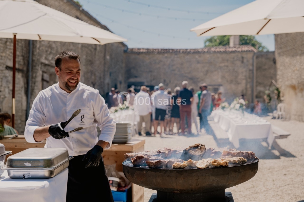 Un chef cuisine des viandes sur un barbecue en plein air lors d'un événement avec des invités en arrière-plan.