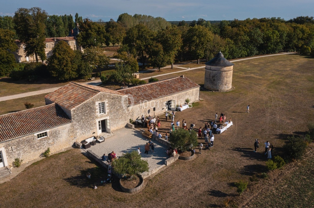 Vue aérienne d'un domaine rural avec bâtiments en pierre, une tour ronde et des personnes réunies en extérieur.