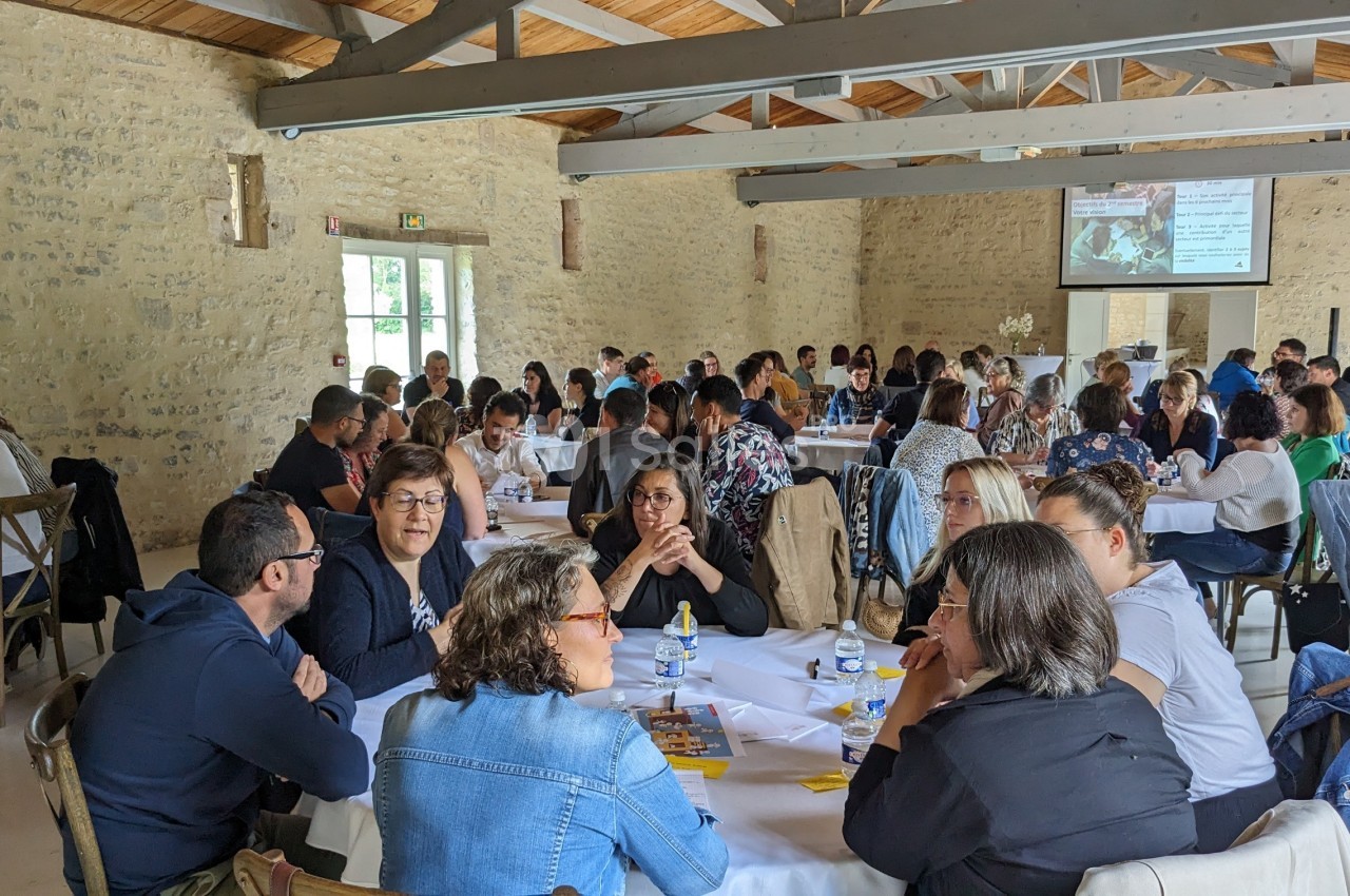 Des personnes assises autour de tables rondes dans une salle lumineuse, participant à une réunion ou un atelier.