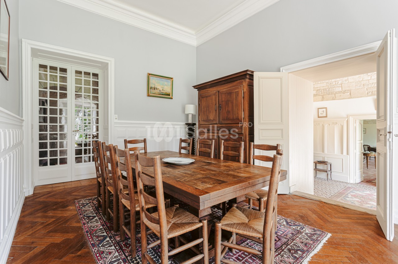 Salle à manger lumineuse avec table en bois, chaises en paille, armoire ancienne et parquet en chevrons.