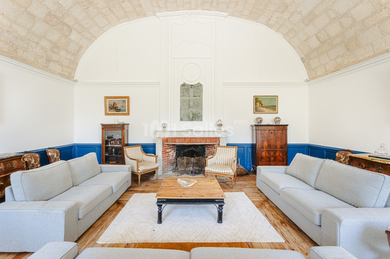 Salon lumineux avec murs blancs, plafond voûté, cheminée centrale, meubles anciens et canapés gris.