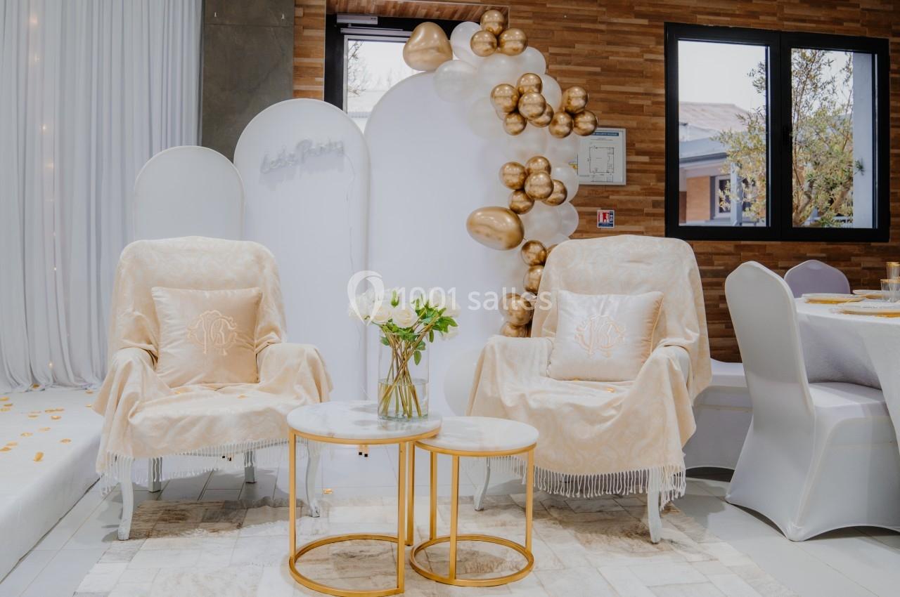 Deux fauteuils beige décorés, entourés de ballons dorés et d'une table basse avec un vase de fleurs blanches.